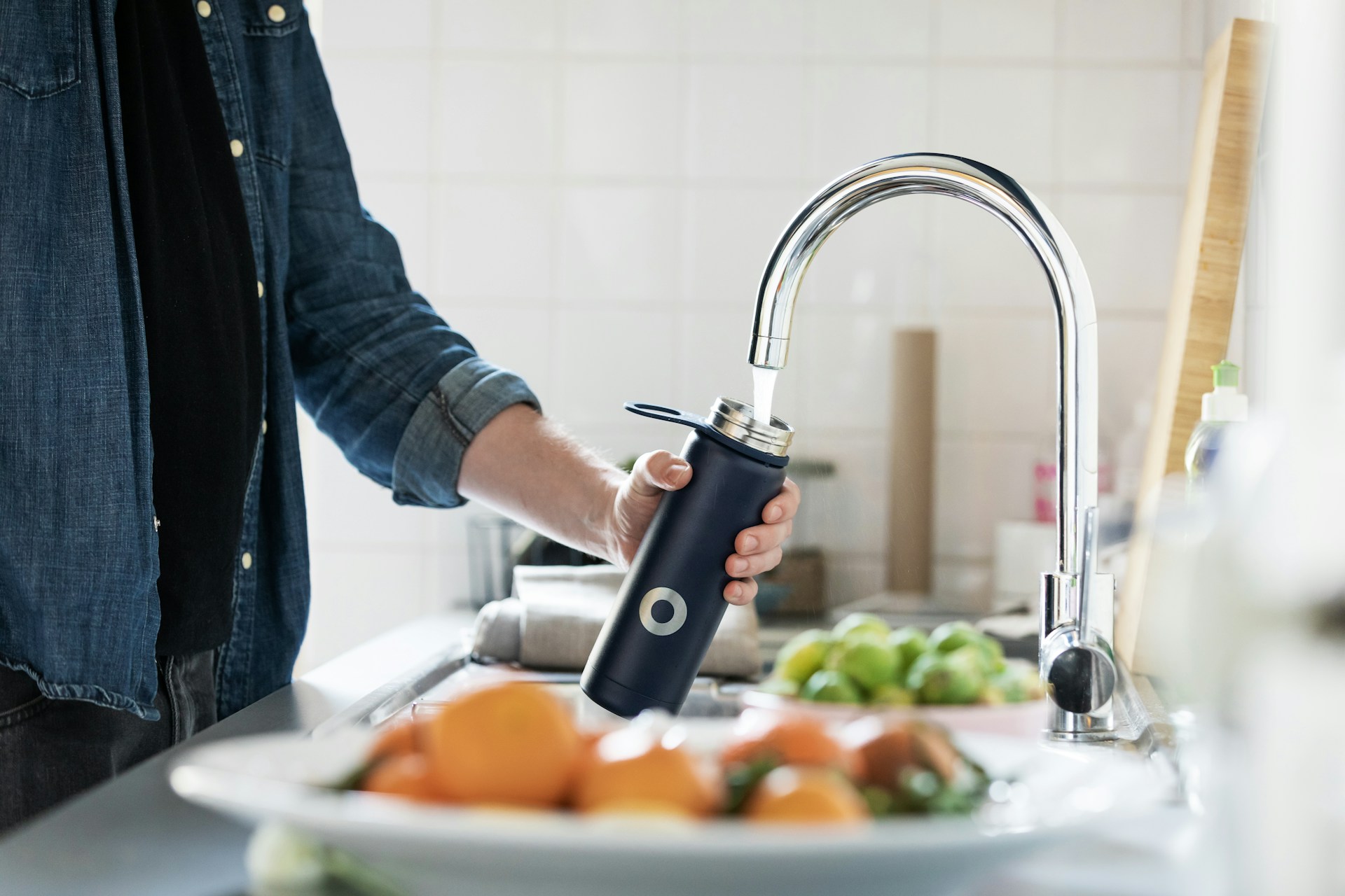 A person filling their water bottle under the sink with a plate of fruit on the counter